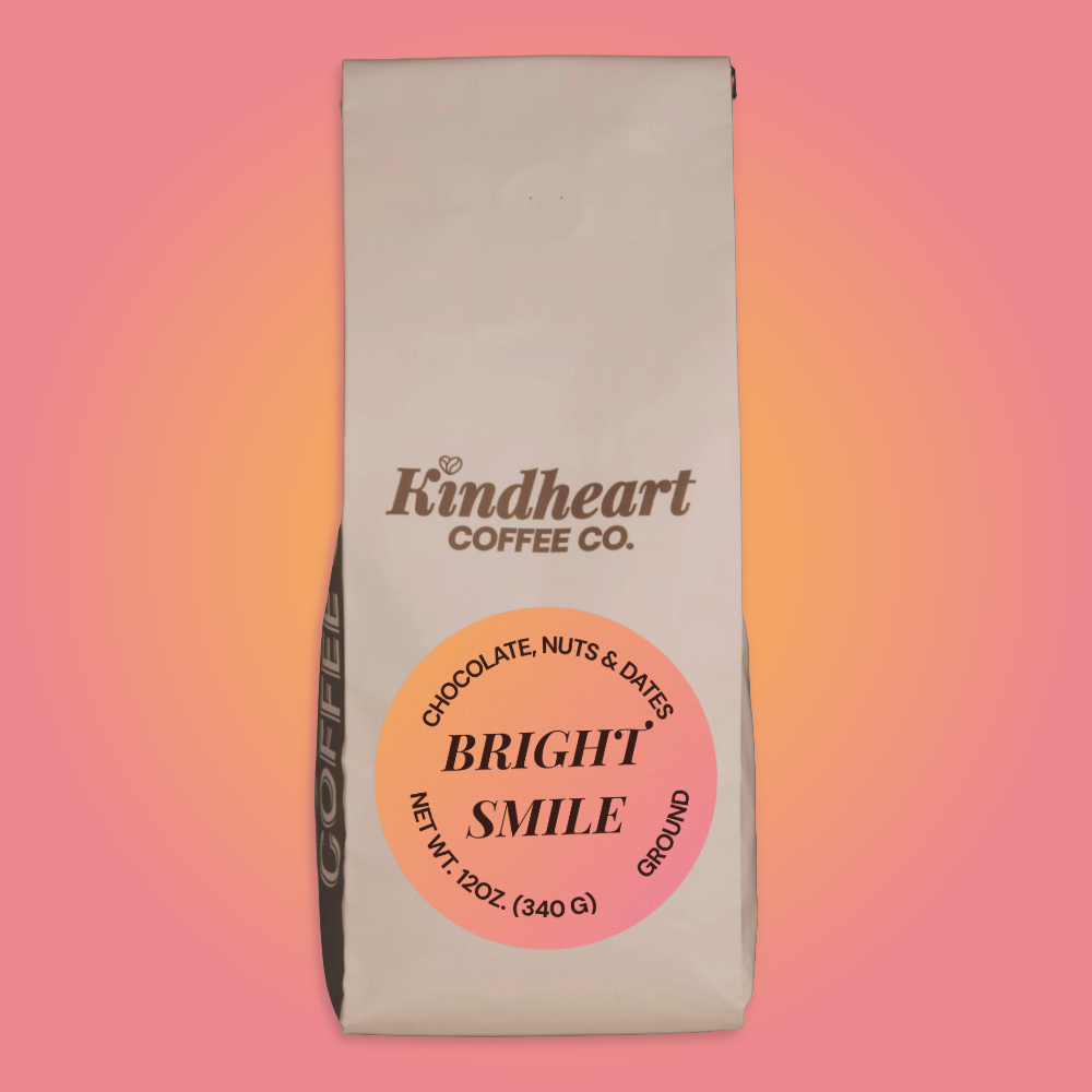 A 12 oz bag of Kindheart Coffee Co. 'Bright Smile' ground coffee, featuring a cream-colored bag with a prominent pink and orange circular label, set against a pink-to-orange gradient background.