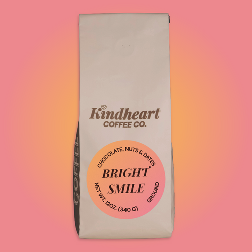 A 12 oz bag of Kindheart Coffee Co. 'Bright Smile' ground coffee, featuring a cream-colored bag with a prominent pink and orange circular label, set against a pink-to-orange gradient background.
