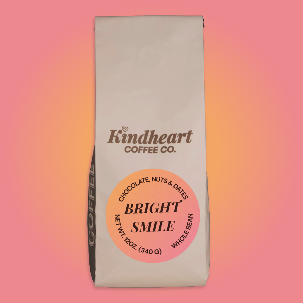 A 12 oz bag of Kindheart Coffee Co. 'Bright Smile' whole bean coffee, featuring a cream-colored bag with a prominent pink and orange circular label, set against a pink-to-orange gradient background.